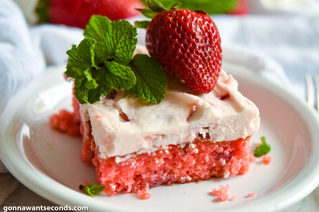 A slice of strawberry jello cake with frosting, topped with fresh strawberry and mint sprigs