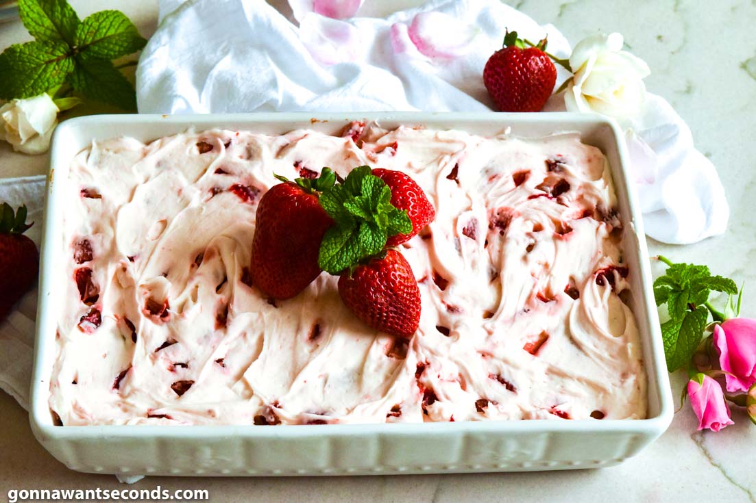 Strawberry jello cake with frosting, topped with fresh strawberries and mint sprigs, in a baking dish