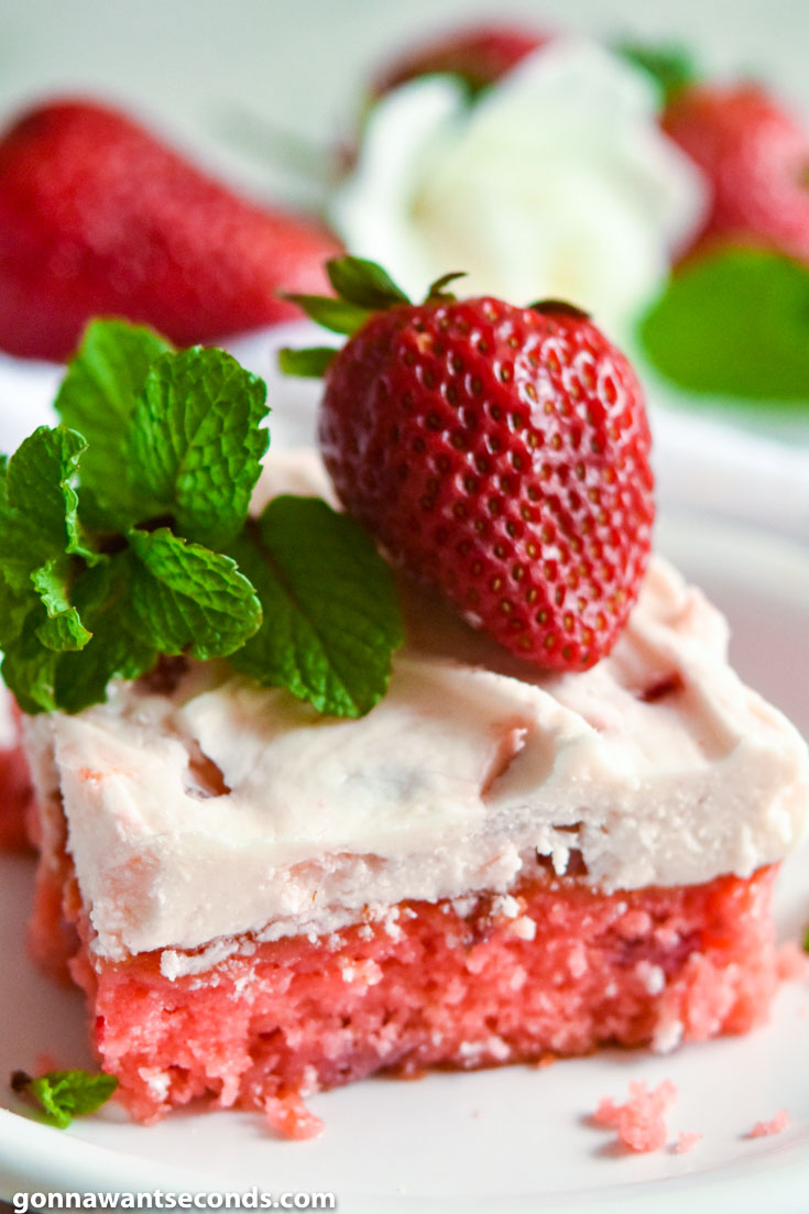 A slice of strawberry jello cake with frosting, topped with fresh strawberry and mint sprigs