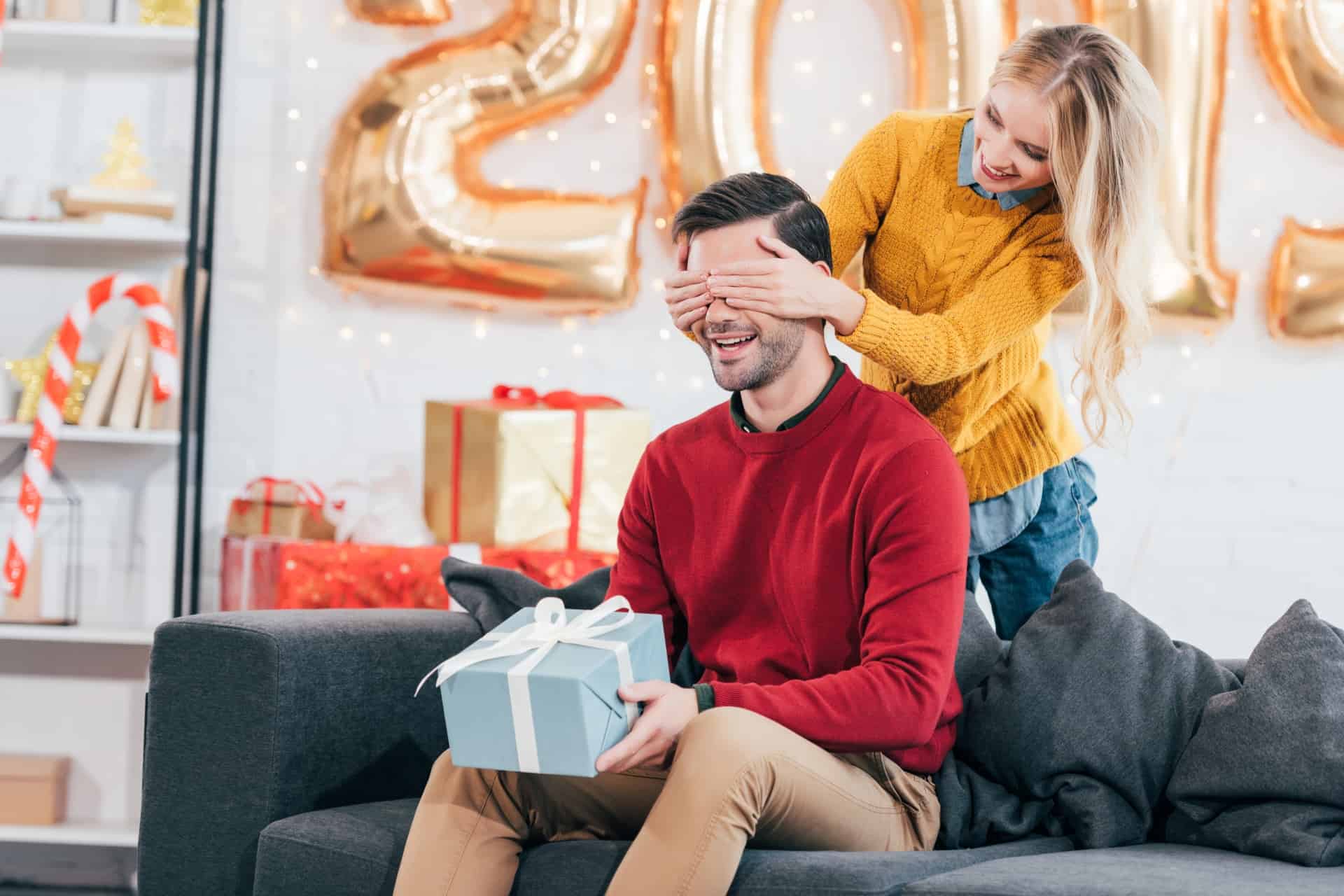 24 Best Christmas Gifts For Your Hubby man holding a gift box while a woman covers his eyes with her hands