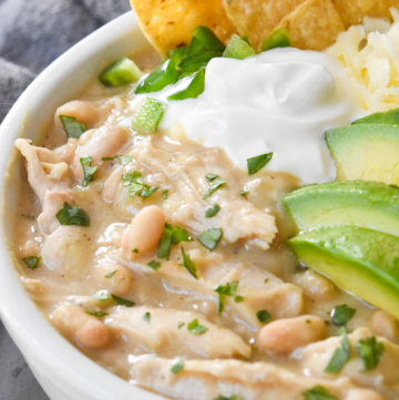 crockpot white chicken chili topped with sliced avocados, cheese, and tortilla chips, in a white bowl