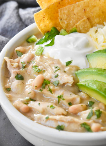 crockpot white chicken chili topped with sliced avocados, cheese, and tortilla chips, in a white bowl