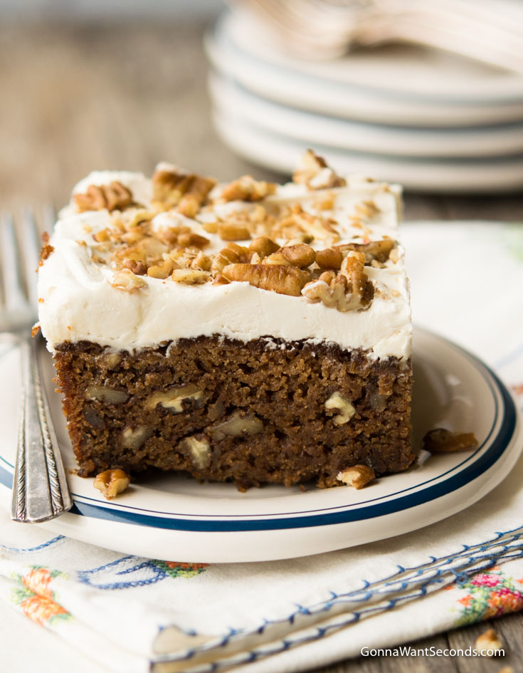 A single serving of Applesauce Cake with frosting and chopped pecans on top, on a saucer 