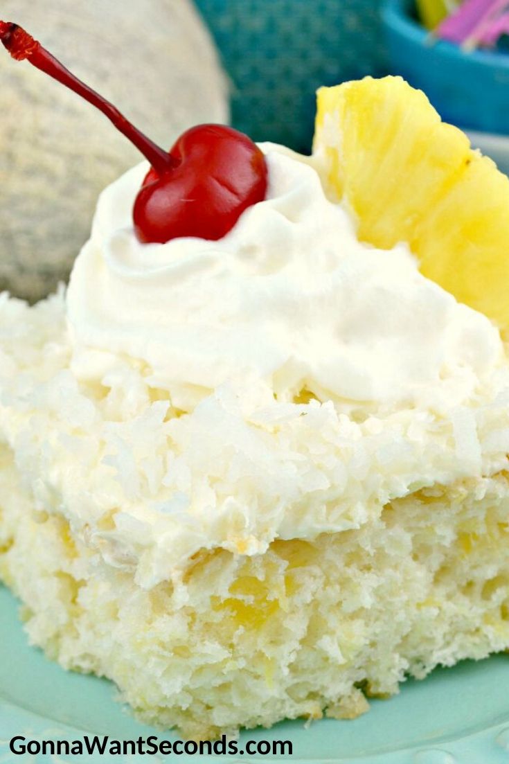 A slice of Pina Colada Cake with frosting, topped with a cherry, a pineapple, and a cocktail umbrella