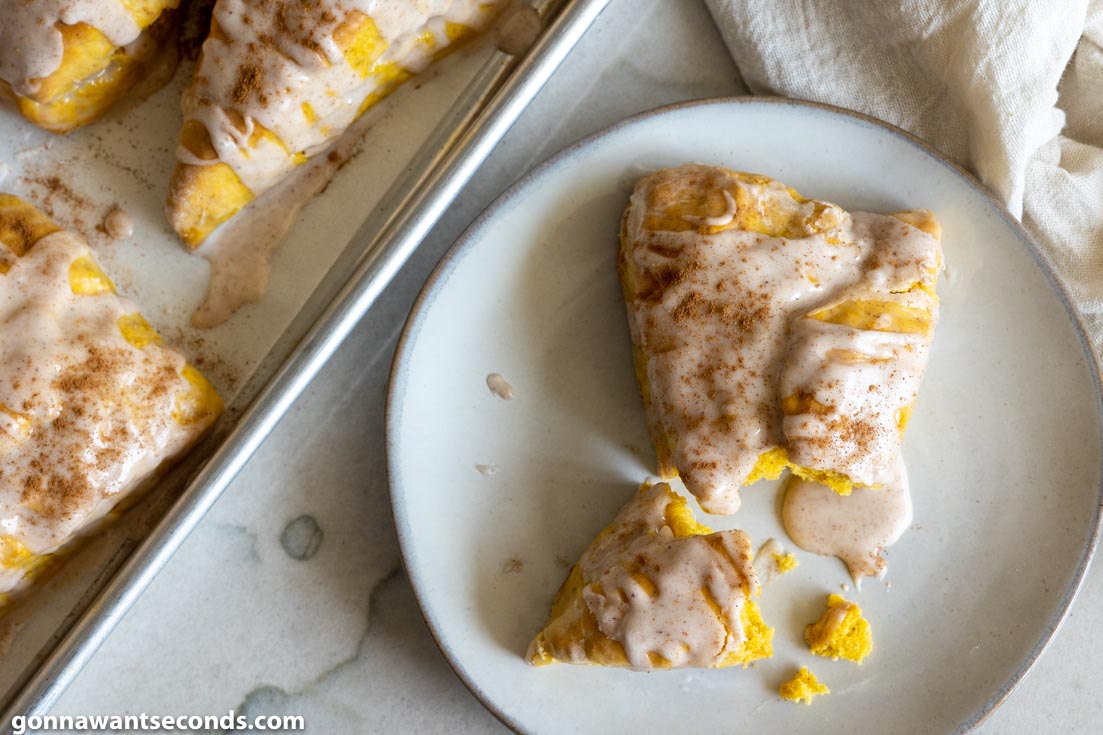 A slice of pumpkin breakfast scones on a plate with a baking sheet full of scones on the side