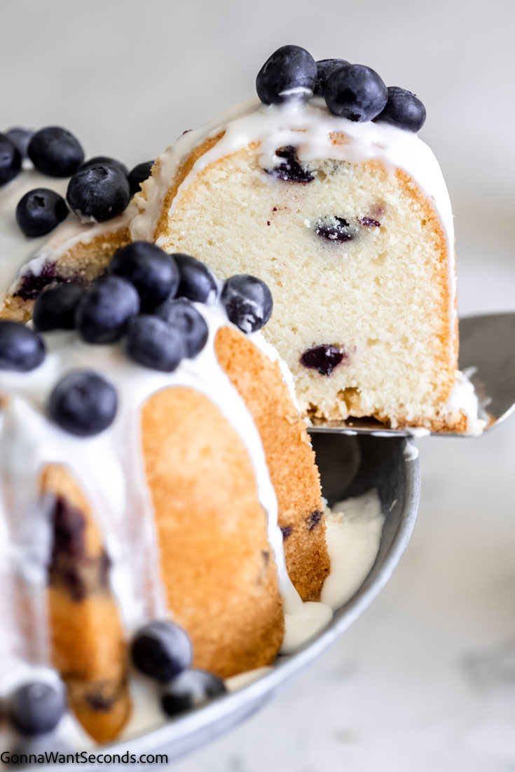 lifting a slice of cake from a blueberry pound cake bundt with glaze topped with fresh blueberries