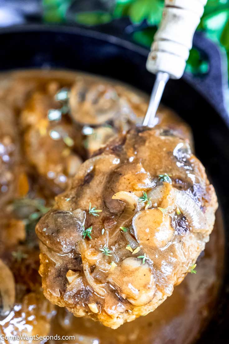 Our salisbury steak is melt-in-your-mouth tender, perfectly seasoned, then smothered in mushroom onion gravy. Just like Grandma's! #hamburgersteak #salisburysteak Close up of ladle scooped a salisbury steak from the pan