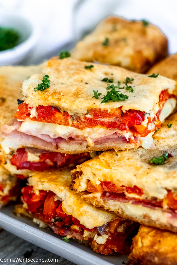 These antipasto squares are incredibly tasty to serve at a football party or any gathering. It has crispy dough, ham, salami, provolone, and Swiss cheese! #antipasto #antipastosquares a pile of antipasto squares on a serving platter