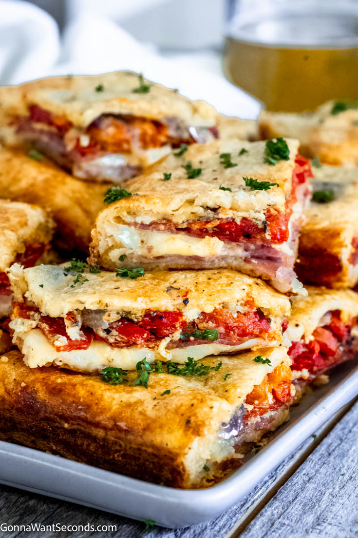 These antipasto squares are incredibly tasty to serve at a football party or any gathering. It has crispy dough, ham, salami, provolone, and Swiss cheese! #antipasto #antipastosquares a pile of antipasto squares on a serving platter