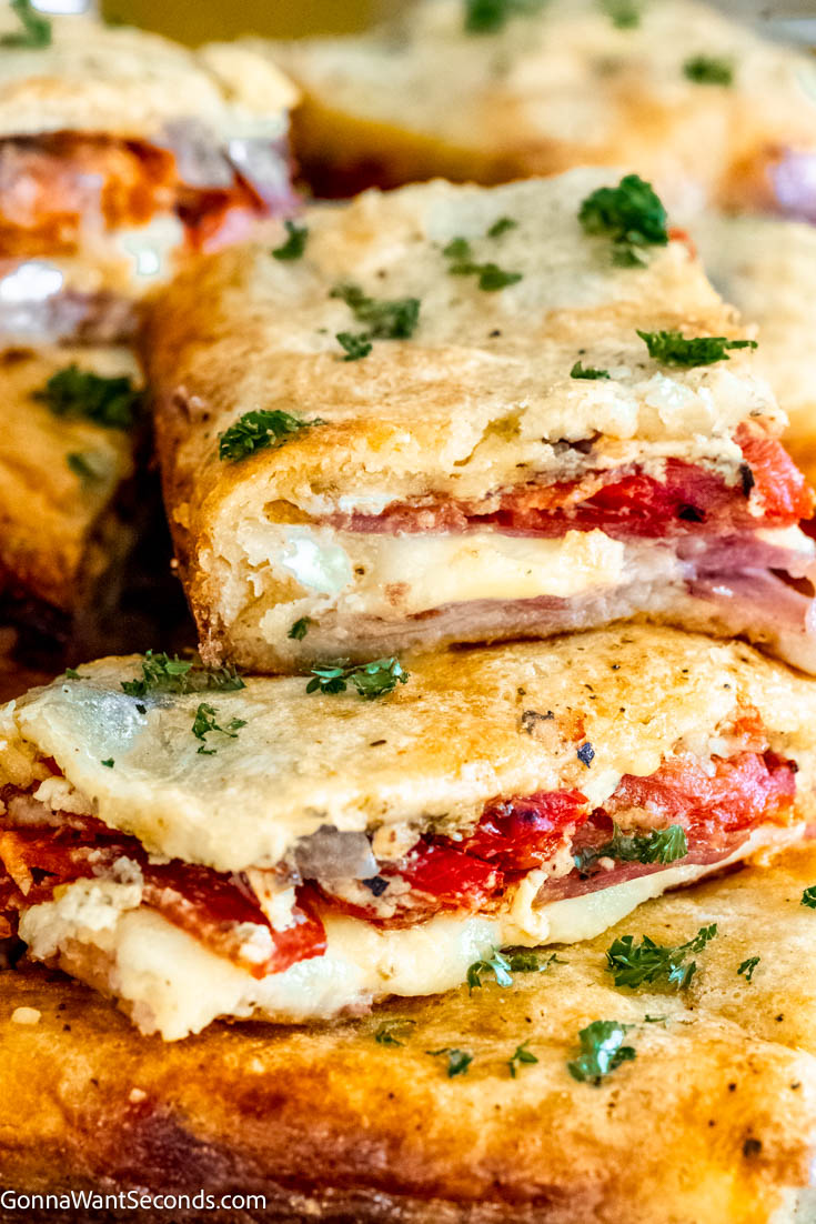 These antipasto squares are incredibly tasty to serve at a football party or any gathering. It has crispy dough, ham, salami, provolone, and Swiss cheese! #antipasto #antipastosquares a pile of antipasto squares on a serving platter