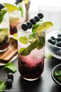 A glass of blueberry mojitos garnished with fresh blueberries on a pick and a wedge of lime.