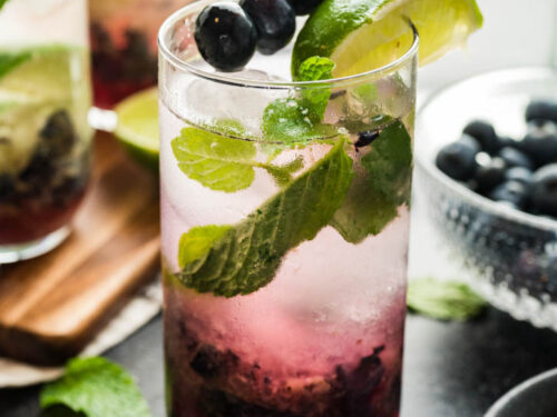 A glass of blueberry mojitos garnished with fresh blueberries on a pick and a wedge of lime.