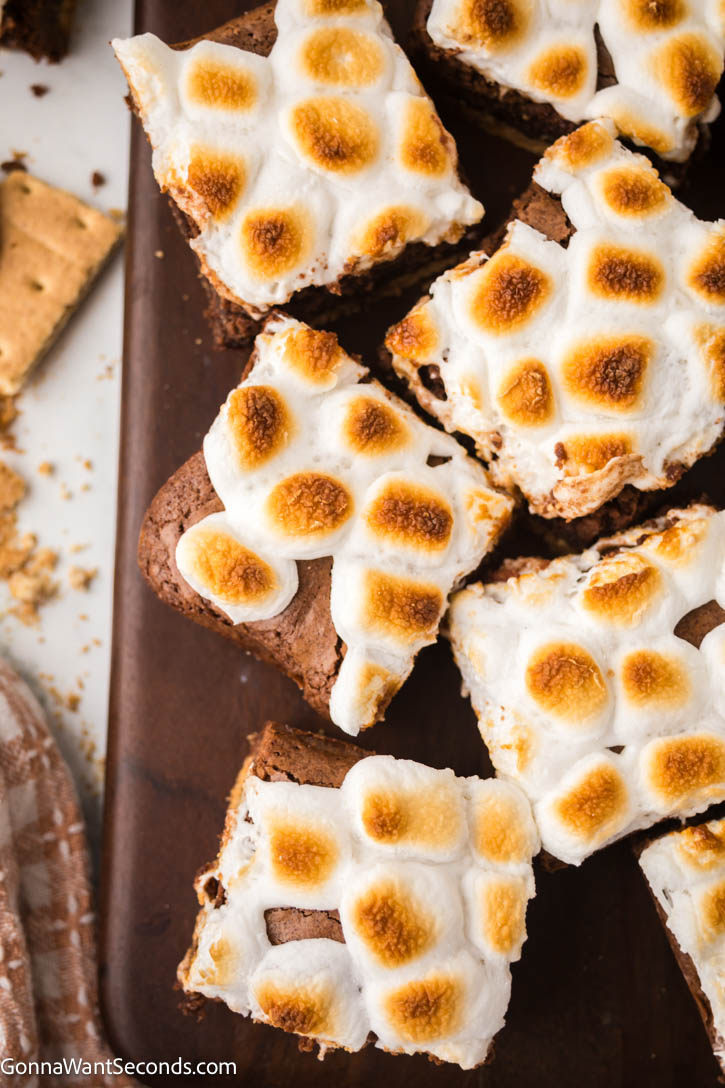 This s'mores brownies has a rich, fudgy brownie base enveloped by a crunchy graham cracker crust, topped off with a layer of ooey-gooey melted marshmallows. #smores #brownies slices of s'mores brownies on a wooden cutting board