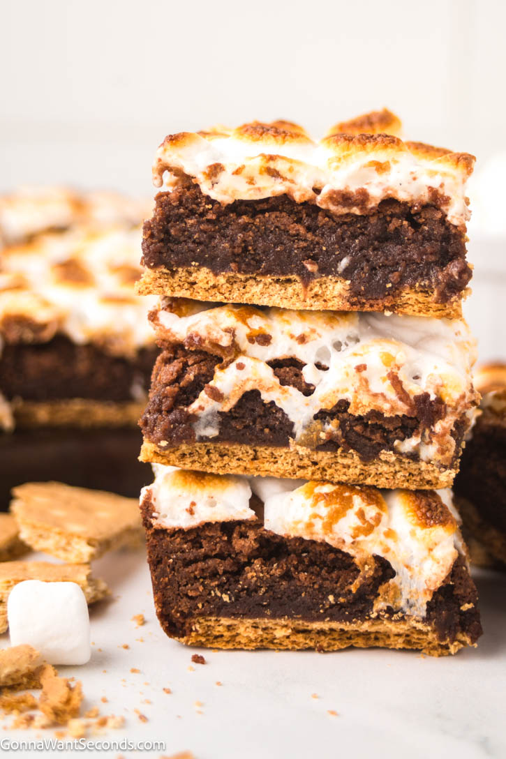 This s'mores brownies has a rich, fudgy brownie base enveloped by a crunchy graham cracker crust, topped off with a layer of ooey-gooey melted marshmallows. #smores #brownies slices of s'mores brownies on top of each other