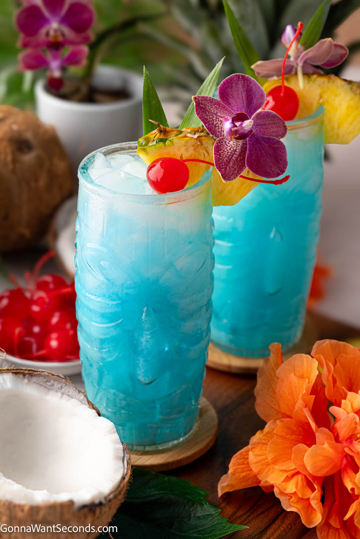 The blue Hawaiian cocktail, a tropical classic with a bright ocean-blue color, creamy coconut, and sweet pineapple flavor, is a delightful vacation in a glass. #bluehawaiiancocktails #cocktails two glasses of Blue Hawaiian Cocktail with an orchid, cherry, and pineapple as garnish