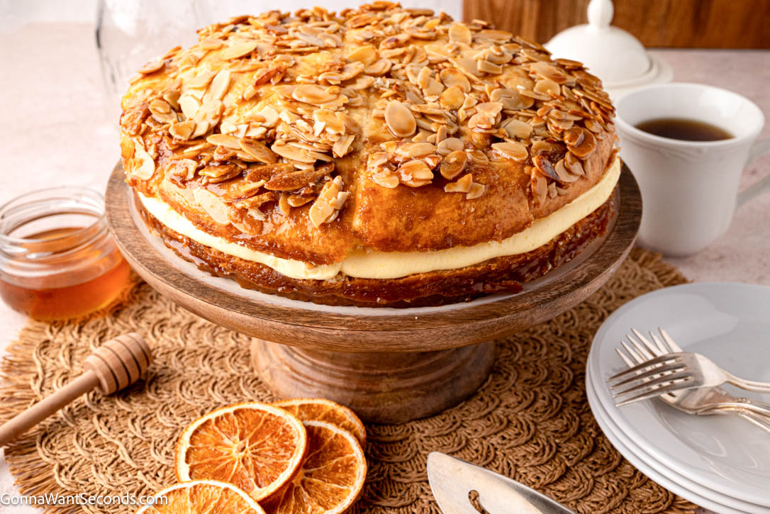 Bee Sting Cake features a soft brioche base, honey-almond topping, and creamy vanilla filling — a dreamy German dessert everyone will love! #beestingcake #bienenstich whole Bee Sting Cake with honey-almond topping and creamy vanilla filling on a wooden cake stand