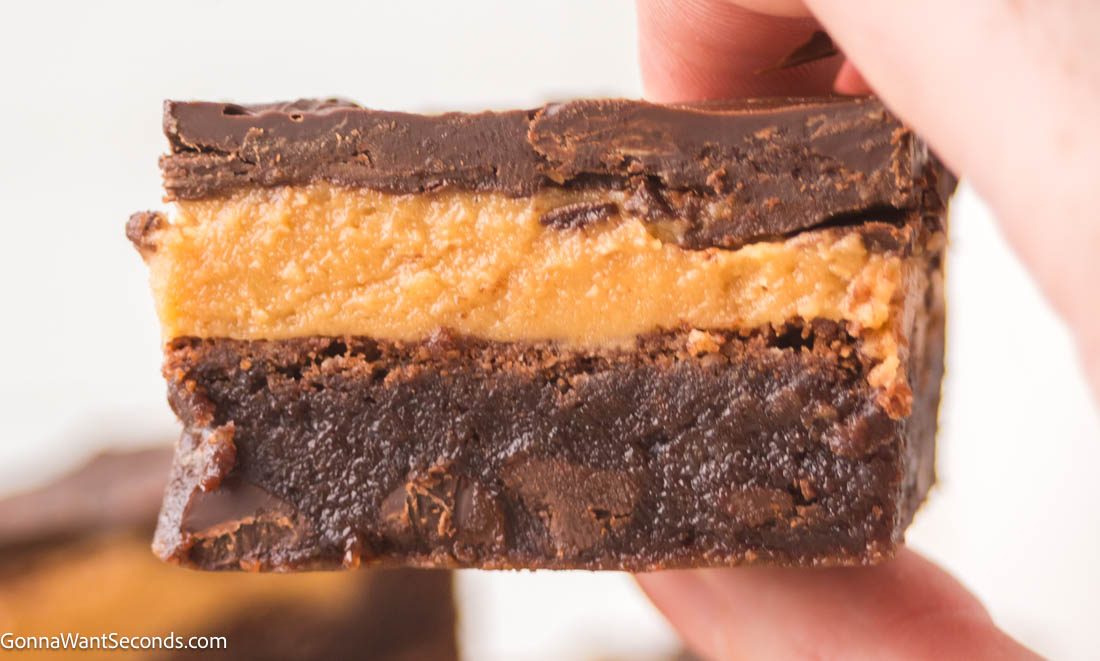Ultra-fudgy Buckeye Brownies with a creamy peanut butter layer + glossy chocolate topping. A Nostalgic chocolate-PB dessert everyone loves. #buckeyerecipes #brownies hand holding a piece Buckeye Brownies with a creamy peanut butter layer and glossy chocolate topping