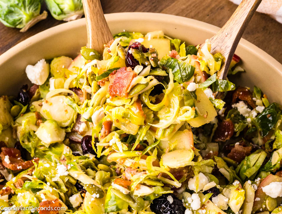 Warm shaved Brussels sprouts salad with apples, bacon, cranberries, and feta in a maple mustard dressing — cozy, colorful, and ready in 20 minutes! #Brusselssprout #Brusselssproutsalad tossing shaved Brussels sprouts salad with apples, bacon, cranberries, and feta