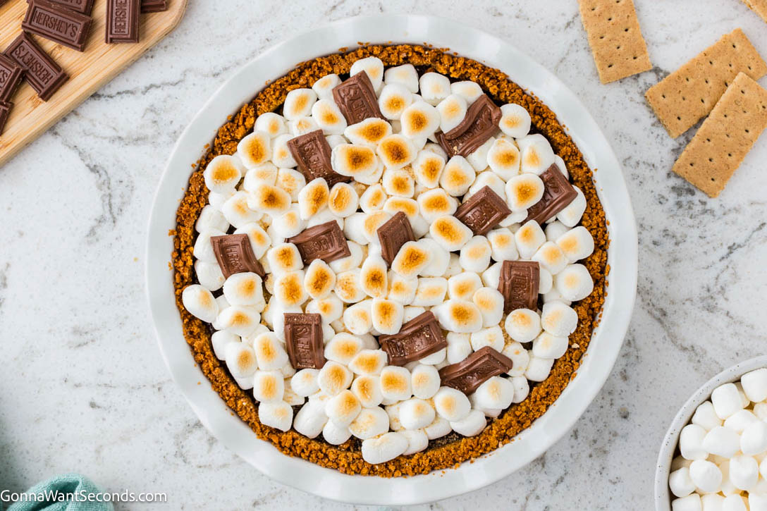 S’mores Pie has a buttery graham crust, silky chocolate filling, and golden toasted marshmallows—gooey, sliceable, and simply irresistible! #smores #smorespie whole s'mores pie with toasted marshmallows and small hershey chocolate bars on top