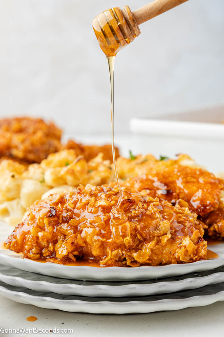 These Hot Honey Chicken Tenders are baked in the oven with a crunchy cornflake coating and finished with a sticky-sweet, lightly spicy hot honey glaze. #chickentenders #hothoneychicken Drizzling honey over Hot Honey Chicken Tenders