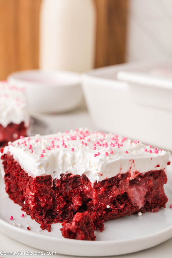 Ultra-moist red velvet poke cake filled with creamy cheesecake pudding and topped with fluffy whipped topping — an easy, make-ahead dessert! #redvelvetcake #pokecake a slice of Red velvet poke cake with whipped topping and pink sprinkles