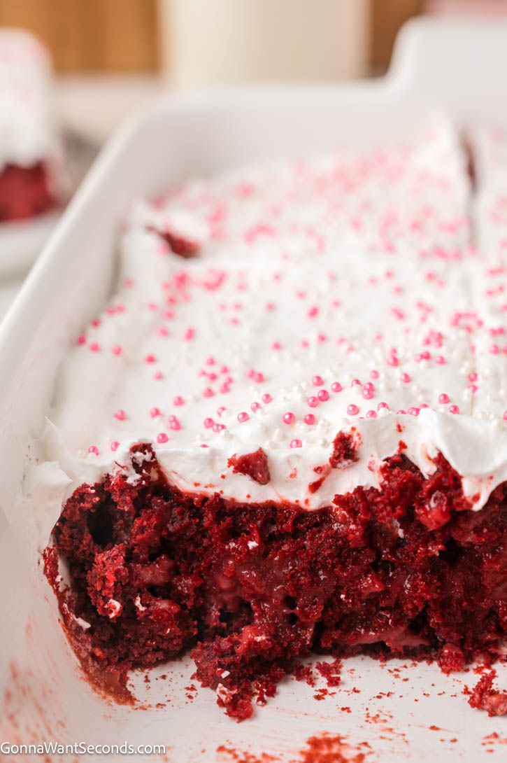 Ultra-moist red velvet poke cake filled with creamy cheesecake pudding and topped with fluffy whipped topping — an easy, make-ahead dessert! #redvelvetcake #pokecake whole Red velvet poke cake with whipped topping and pink sprinkles with a slice taken out
