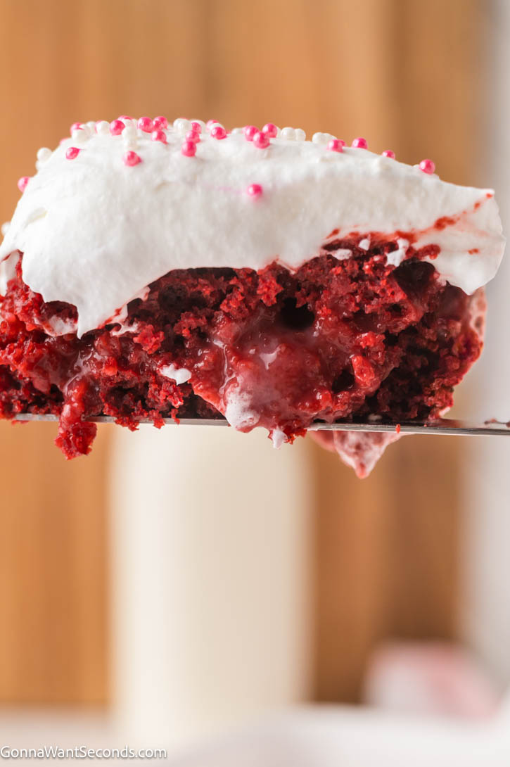 Ultra-moist red velvet poke cake filled with creamy cheesecake pudding and topped with fluffy whipped topping — an easy, make-ahead dessert! #redvelvetcake #pokecake lifting a slice of a slice of Red velvet poke cake with whipped topping and pink sprinkles