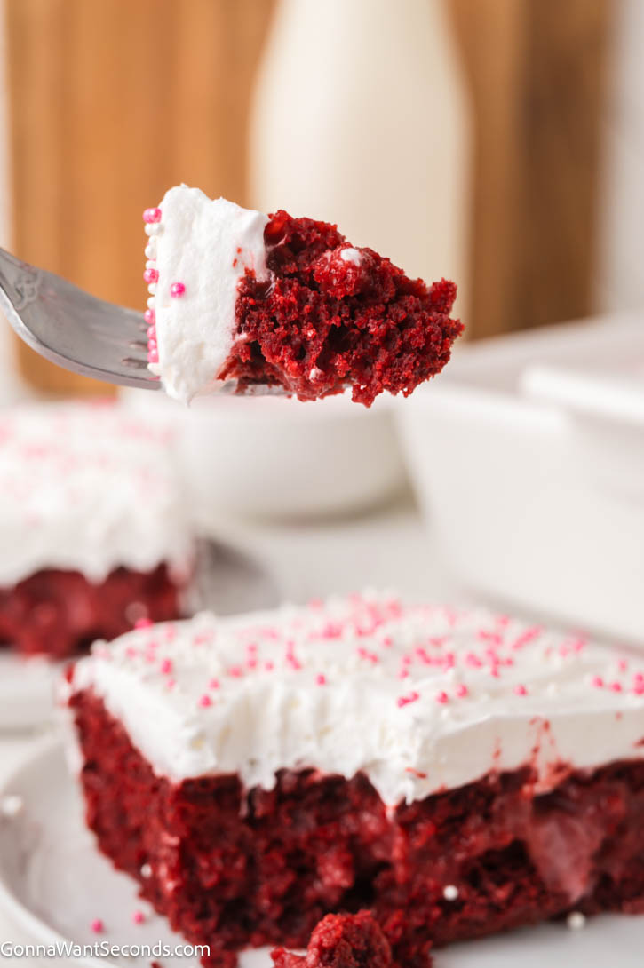 Ultra-moist red velvet poke cake filled with creamy cheesecake pudding and topped with fluffy whipped topping — an easy, make-ahead dessert! #redvelvetcake #pokecake a fork piercing a portion for a slice of a slice of Red velvet poke cake with whipped topping and pink sprinkles