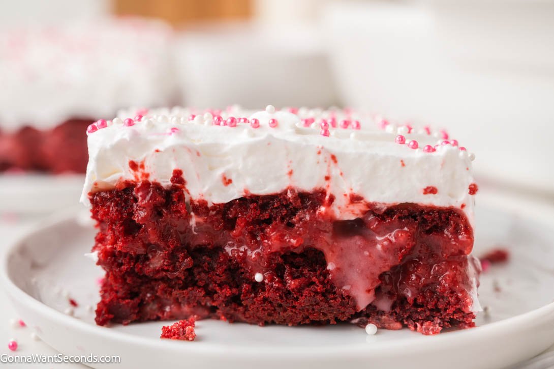 Ultra-moist red velvet poke cake filled with creamy cheesecake pudding and topped with fluffy whipped topping — an easy, make-ahead dessert! #redvelvetcake #pokecake a slice of Red velvet poke cake with whipped topping and pink sprinkles