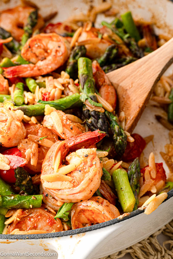 This shrimp orzo recipe is an easy one-pan dinner with juicy shrimp, tender orzo, asparagus, tomatoes, Parmesan, garlic, and fresh lemon. #shrimporzo #shrimprecipes wooden spoon scooping shrimp orzo recipe