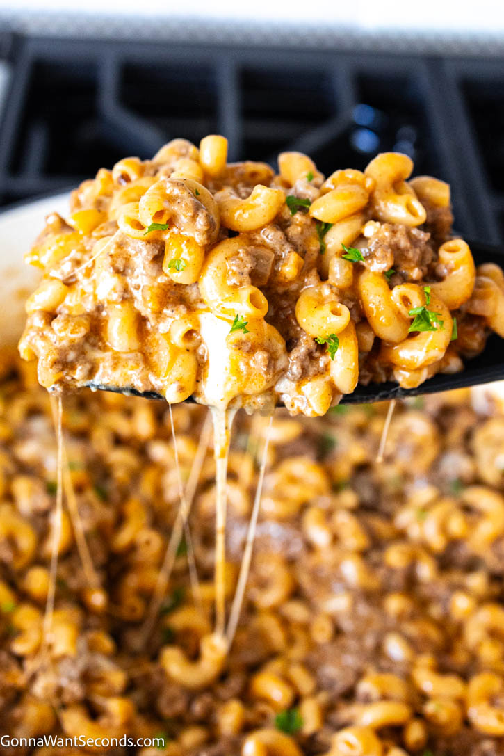 Enjoy the best Homemade Hamburger Helper with tender pasta, seasoned beef, and creamy cheese sauce. This easy one-pan dinner is ready in 50 minutes. #hamburgerhelper #onepandinner Wooden spatula scooping Hamburger helper with cheese strings drips on the pot