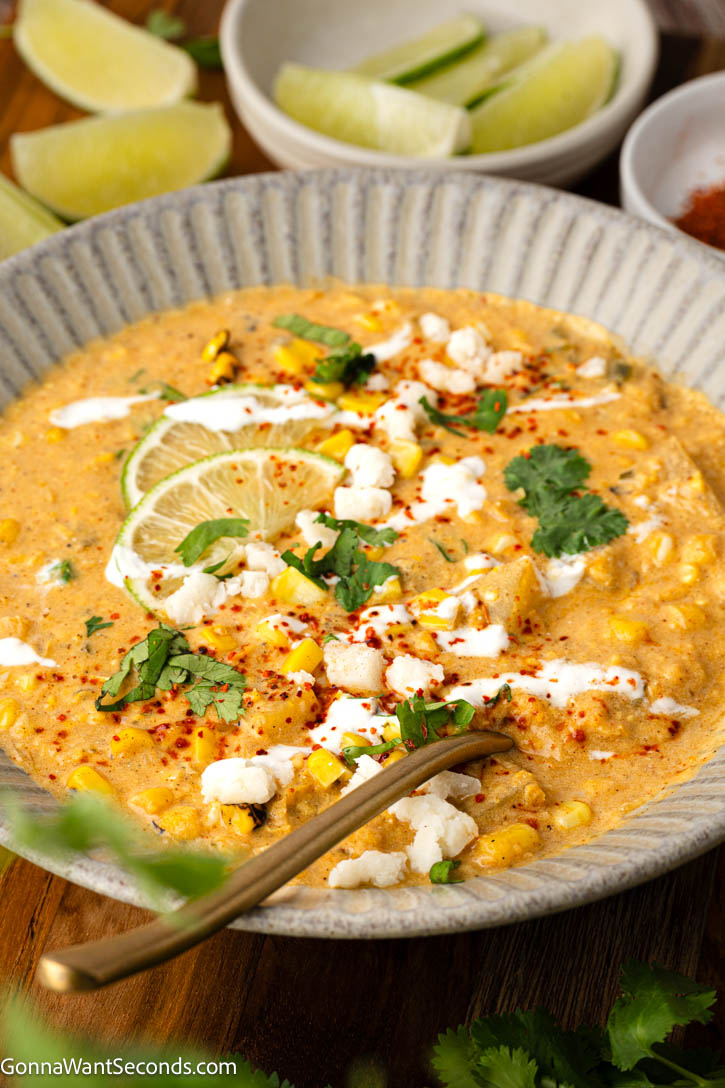 This Mexican Street Corn Soup is rich and creamy with sweet corn, smoky poblano, tender potatoes, lime, cotija, and all your favorite elote flavors. #mexicansoup #streetcornsoup Mexican Street Corn soup topped with lime wedges, cilantro, sour cream and heavy cream in a bowl
