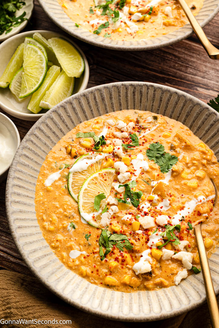 This Mexican Street Corn Soup is rich and creamy with sweet corn, smoky poblano, tender potatoes, lime, cotija, and all your favorite elote flavors. #mexicansoup #streetcornsoup Mexican Street Corn soup topped with lime wedges, cilantro, sour cream and heavy cream in a bowl