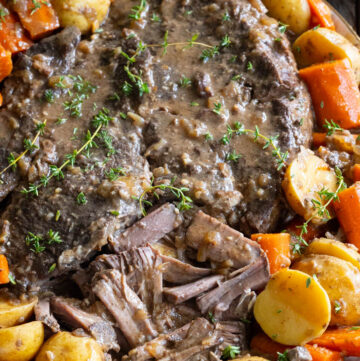 Tender Crockpot pot roast served on a platter surrounded by cooked potatoes, sliced carrots, and rich savory gravy sprigged with fresh thyme.